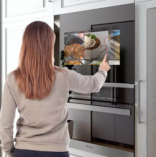 A woman is pointing at a touch-screen kitchen TV built-in above an oven.