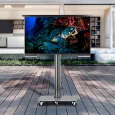 Outdoor TV in wheel-based floor mount in a patio with a white furnished house in the background.