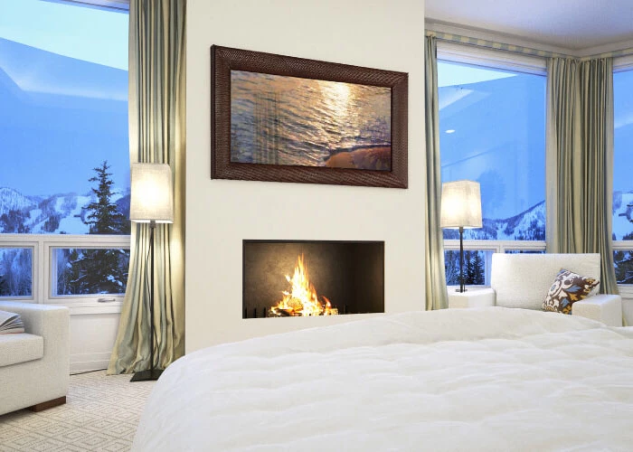 Un dormitorio con un TV espejo enmarcado colocado en una pared blanca sobre una chimenea y ventanas de vidrio amplias.