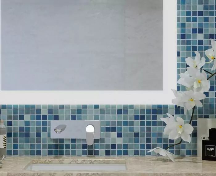 Frosted vanity mirror mounted on a blue mosaic tiled wall above a sink and an orchid in Abu Dhabi.