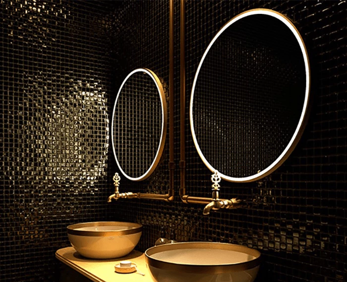 Black mosaic tiled wall with a pair of gold oval lit mirrors, two gold sinks, and faucets in Ajman.