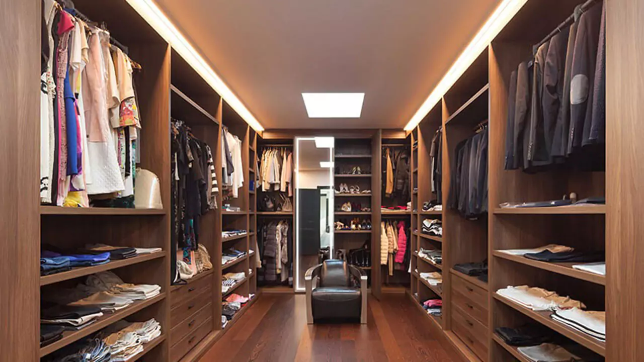Wooden walk in wardrobe with cabinets full of hung and folded clothes and a lighted mirror in Dubai.