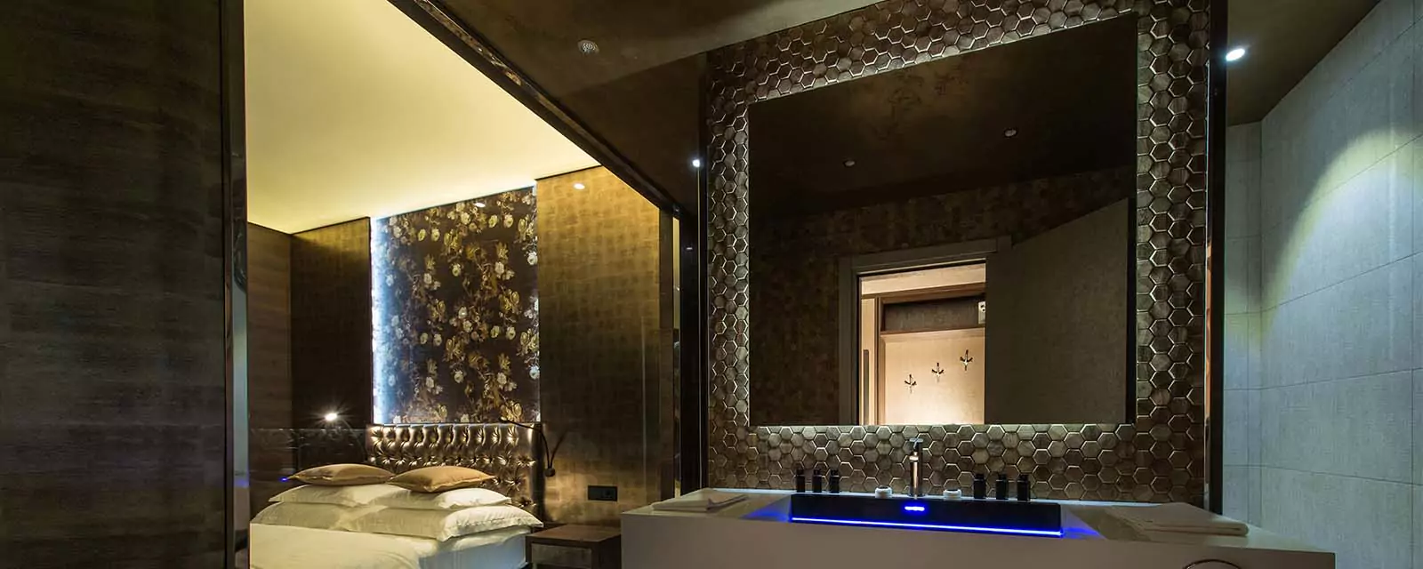 Above a single sink, on a wall with dark hexagon tiles, are Backlit Grand Mirrors from Oman.