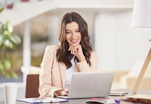 Woman in a beige blazer smiling while looking at a laptop, in a bright office in Kuwait.