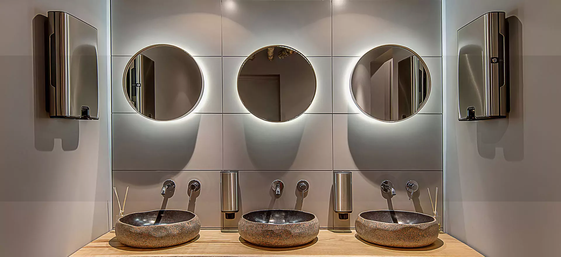 Above the three bathroom sinks, a brown wall is adorned with three circular mirrors from Oman.