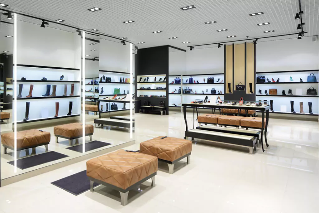 Well lit classy shoe and bag store in Oman with lighted mirrors, leather stools, and a wooden table.