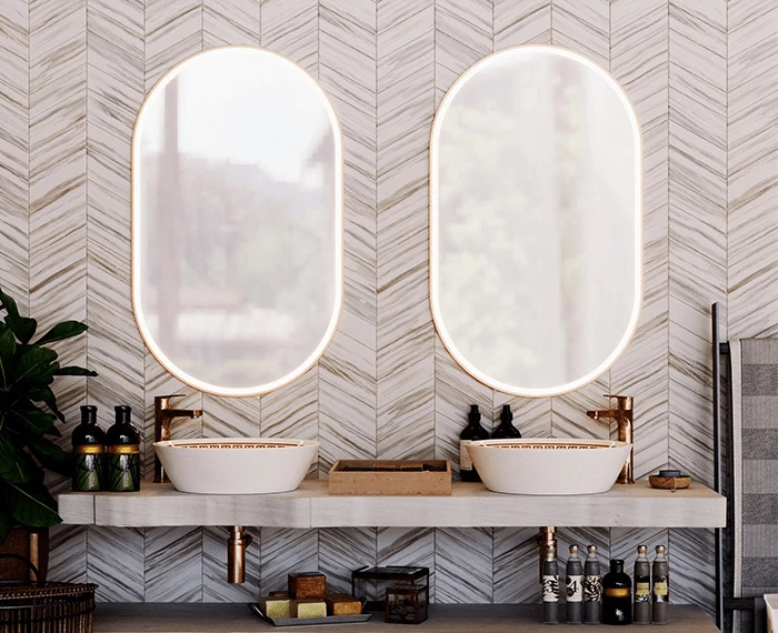 Two oval lighted mirrors attached to a herringbone patterned wall and a dual sink vanity in Sharjah.