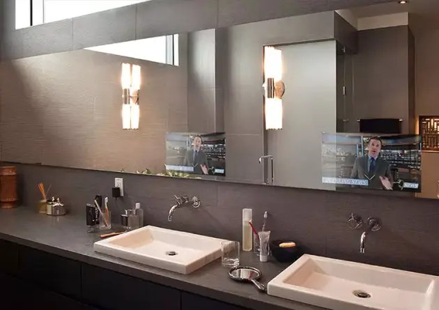 Televisor con espejo ampliado que muestra a un presentador de noticias instalado en una pared marrón sobre un lavabo doble.