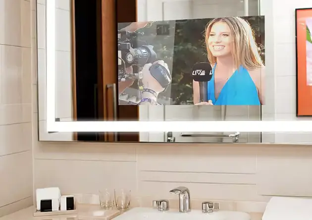 Una alegre presentadora de noticias en un espejo iluminado con pantalla de televisión sobre el lavabo individual.