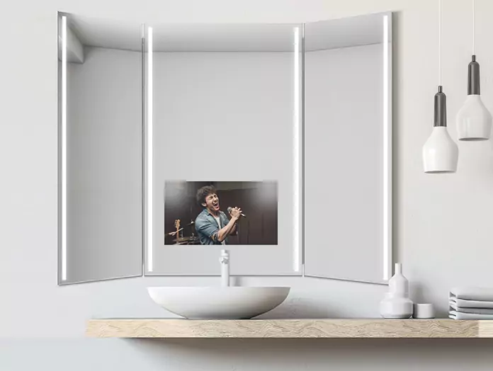 A man singing is displayed on a modern, lighted trifold mirror TV mounted above a basin.