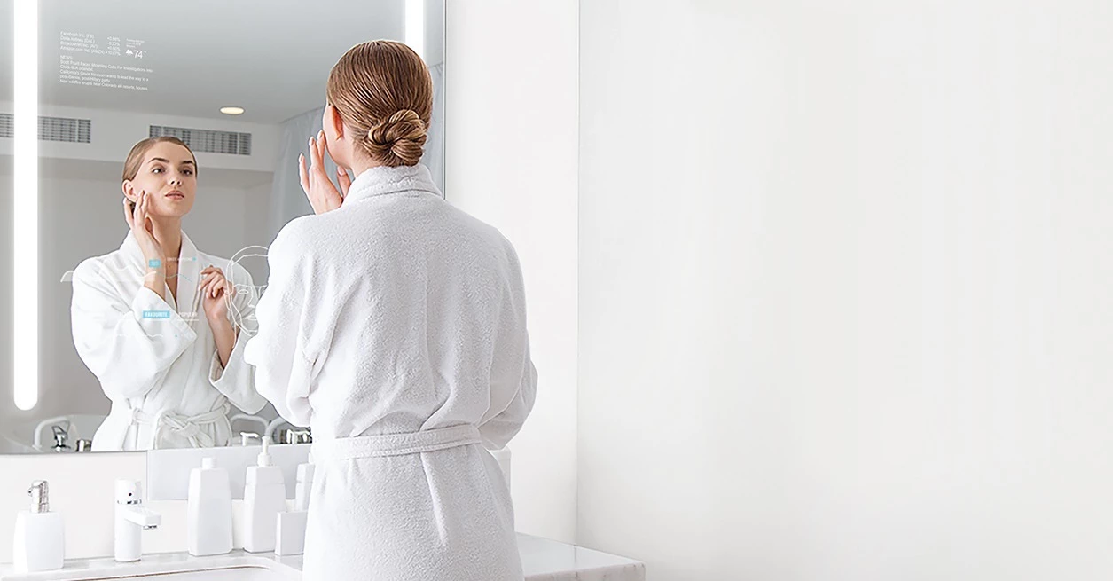 A woman in a bathrobe admiring her reflection in a lighted mirror.