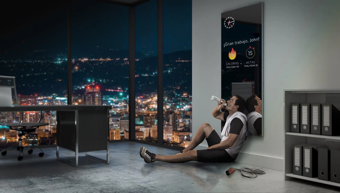 A man sits by a smart fitness mirror showing workout information at the screen at night.