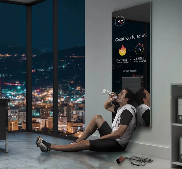 A man is sitting in front of a smart fitness mirror displaying workout statistics during nightime.